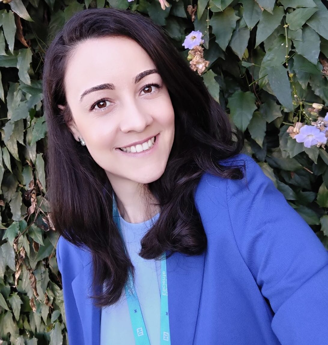 Imagem de Aline Zamoro, ela está com cabelo solto, escuro, e sorridente, usando um casaco azul. Atras dela algumas folhas verdes